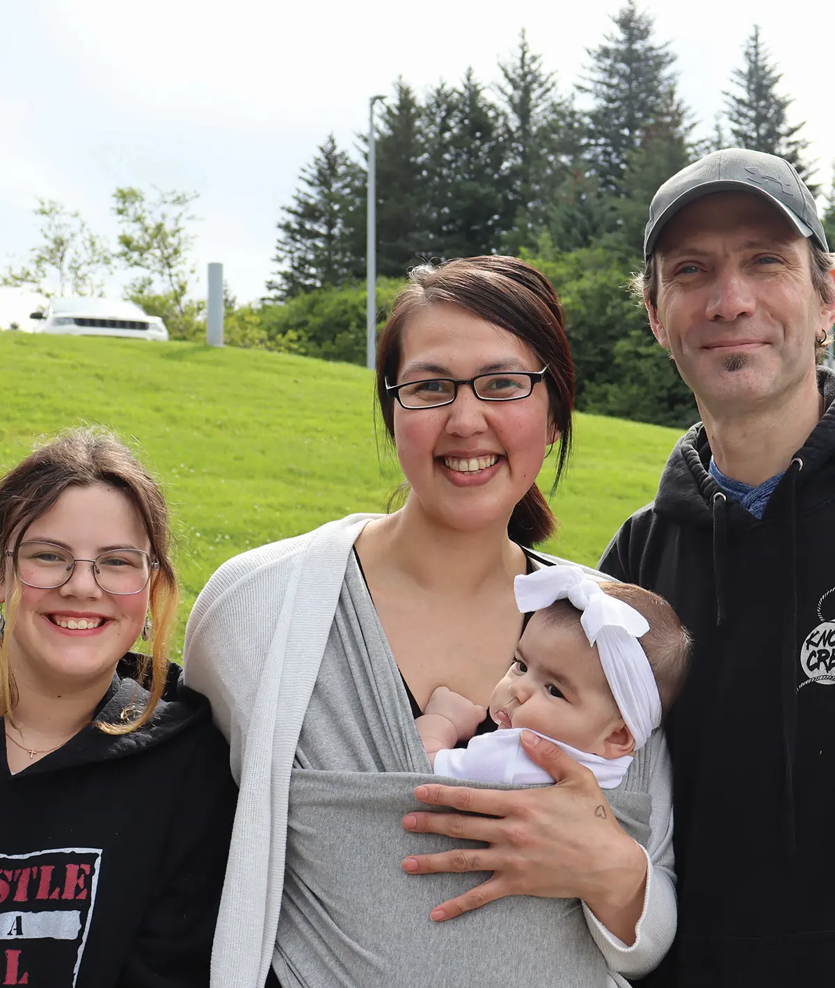 A diverse family of four posing outdoors on a sunny day, with the mother carrying a baby in a soft gray wrap and standing next to her daughter and partner