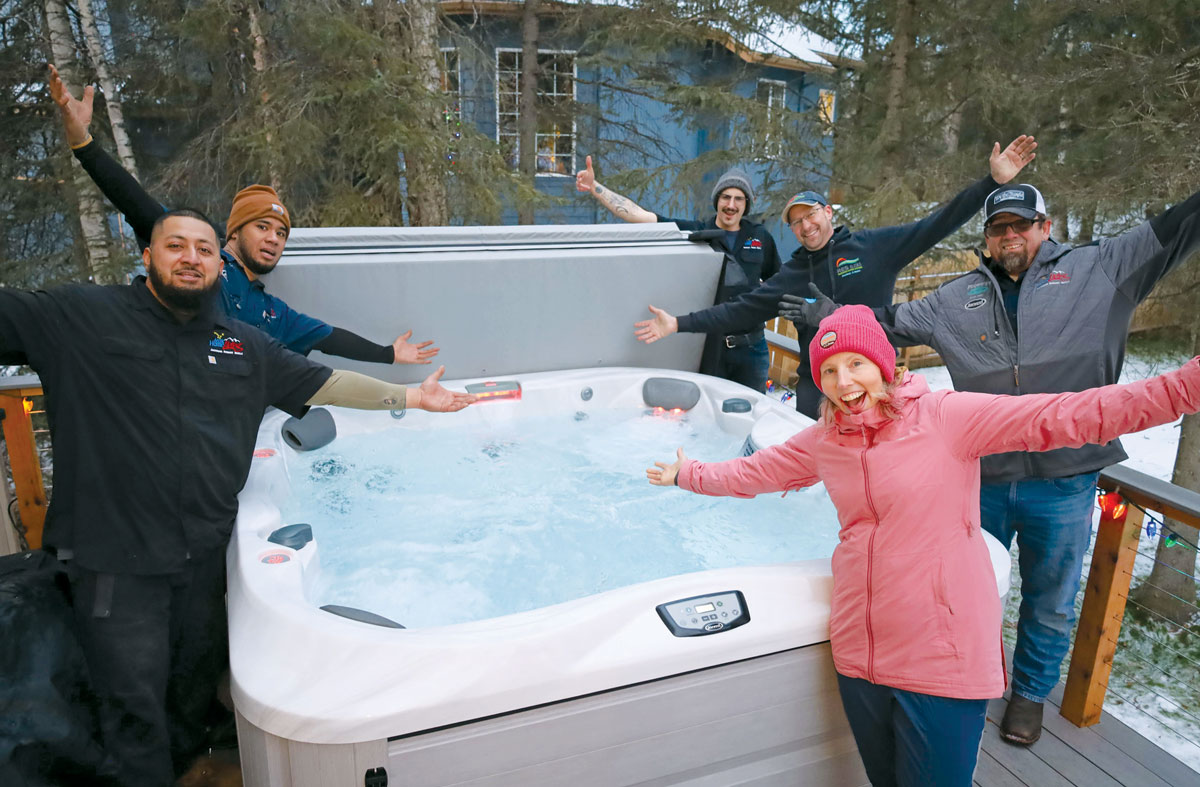 Seven smiling people, including staff and a customer, happily posing around a new outdoor Jacuzzi hot tub on a deck.
