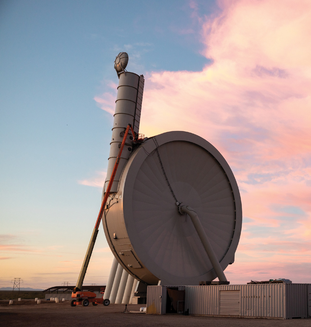 Large, unique industrial structure resembling a massive gray vertical fan, with a worker on a crane lift at sunset.