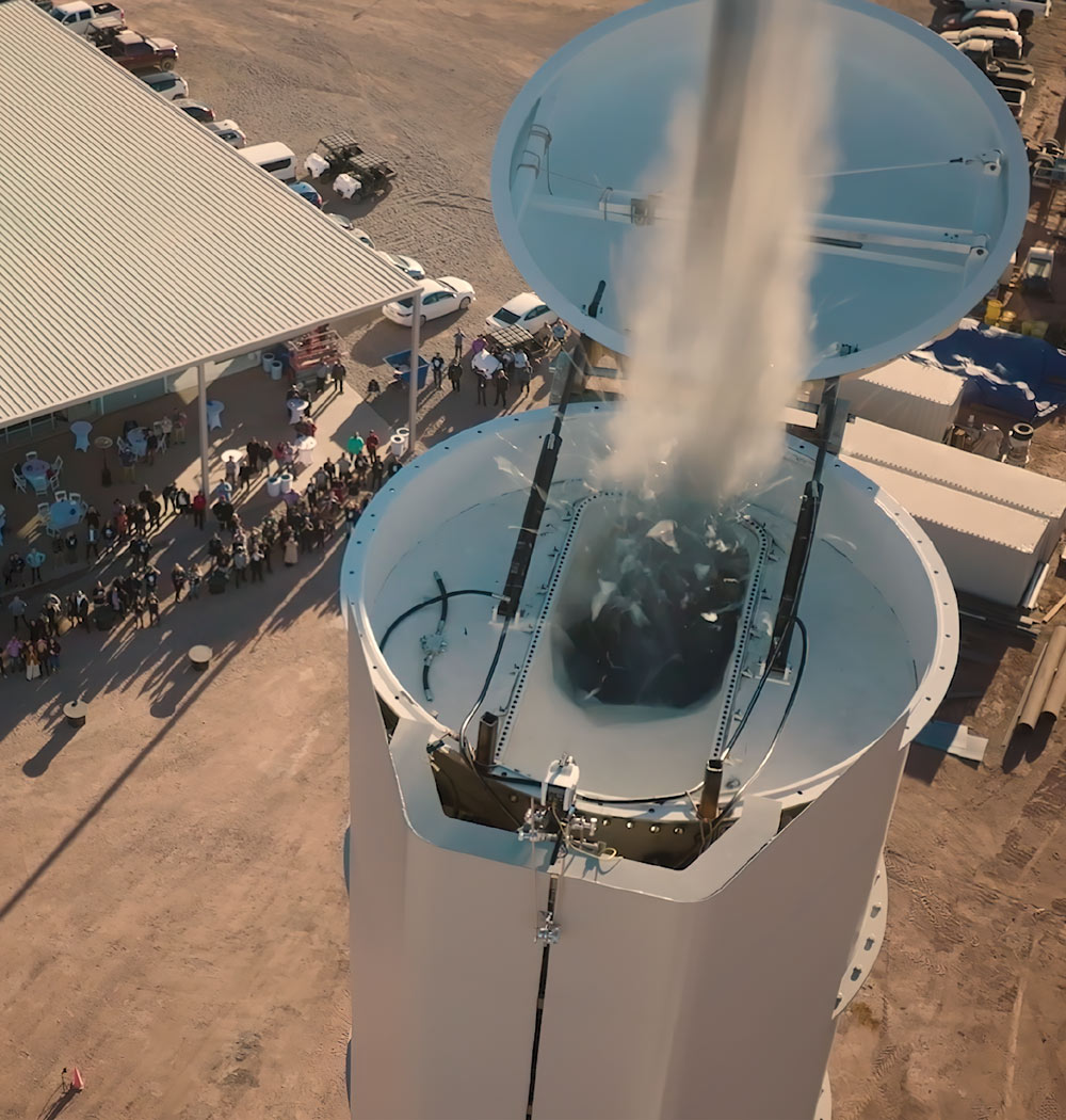 Aerial view of a crowd gathered around a large, tall container structure with its lid open, releasing a cloud of white vapor.