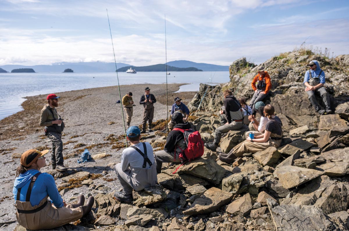 A group of people wearing waders and fishing gear are seated on a rocky Alaska coastline with the ocean and islands visible in the background.