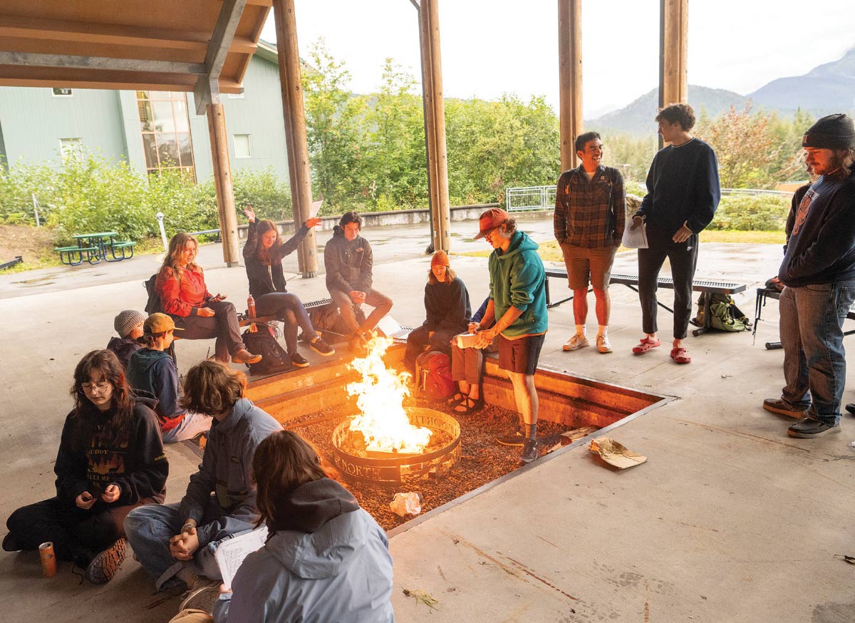 A group of young adults gathered around a large outdoor fire pit under a covered pavilion in a natural, mountainous setting.