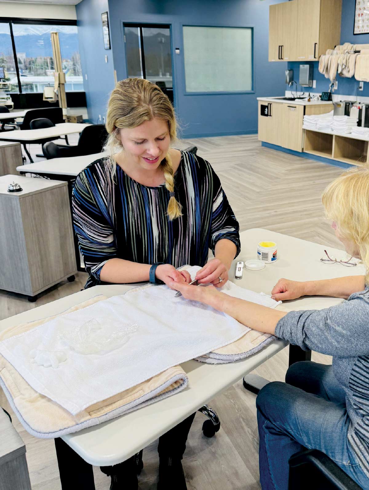 A smiling woman performing a manicure or hand care procedure on an older woman in a brightly lit, modern classroom setting.