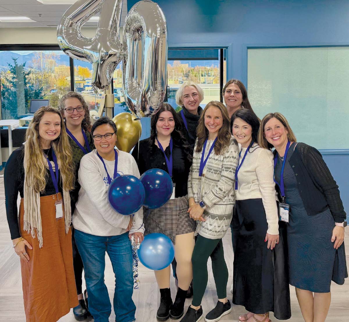 Group photo of nine smiling women standing near large silver "40" balloons and smaller blue and gold balloons in a bright office.