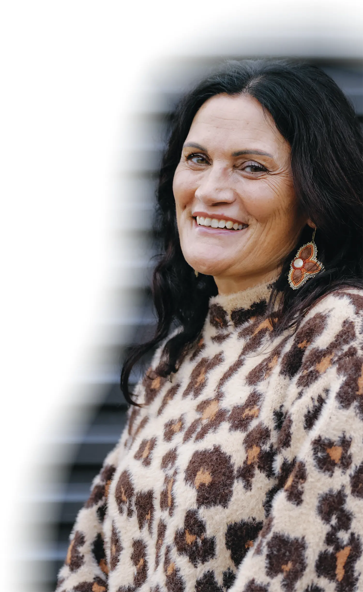 Dena Sommer-Pedebone, a smiling woman with black hair is wearing a fuzzy, brown and tan leopard print sweater and large beaded dangle earrings; The photo is cropped and has a blurred white fade effect along the left edge
