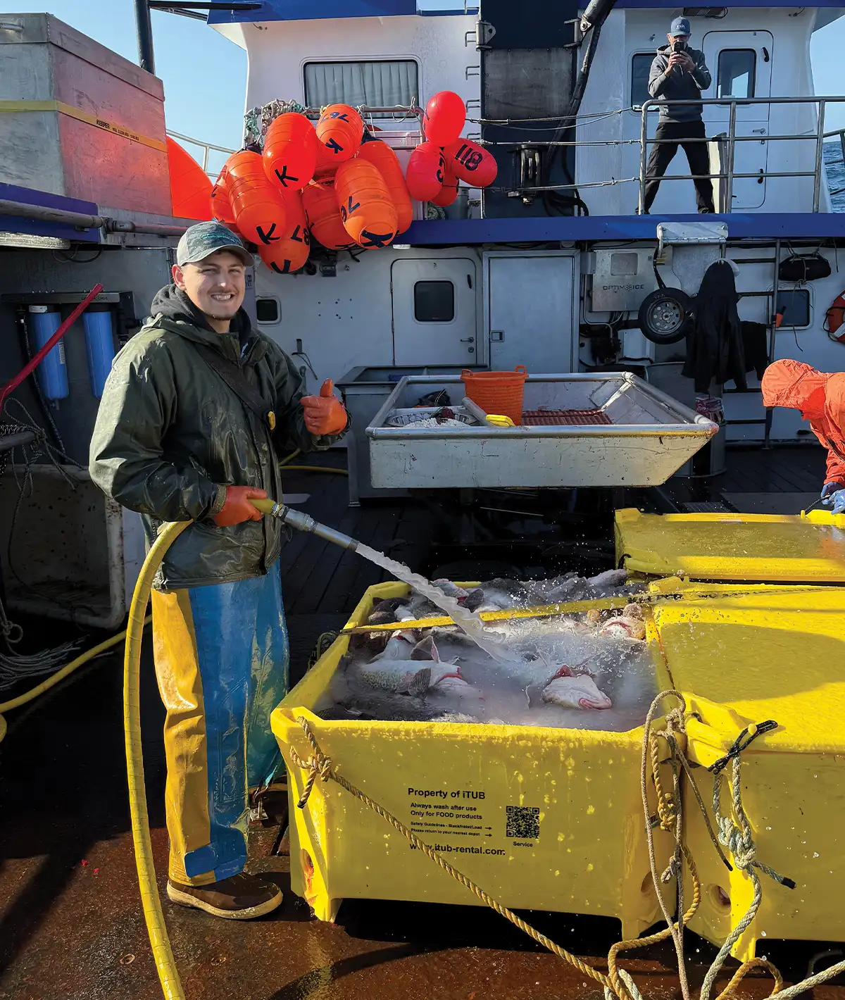A smiling fisherman in waterproof gear sprays water into a large yellow bin filled with fresh fish on the deck of a commercial vessel. Bright orange buoys hang in the background.