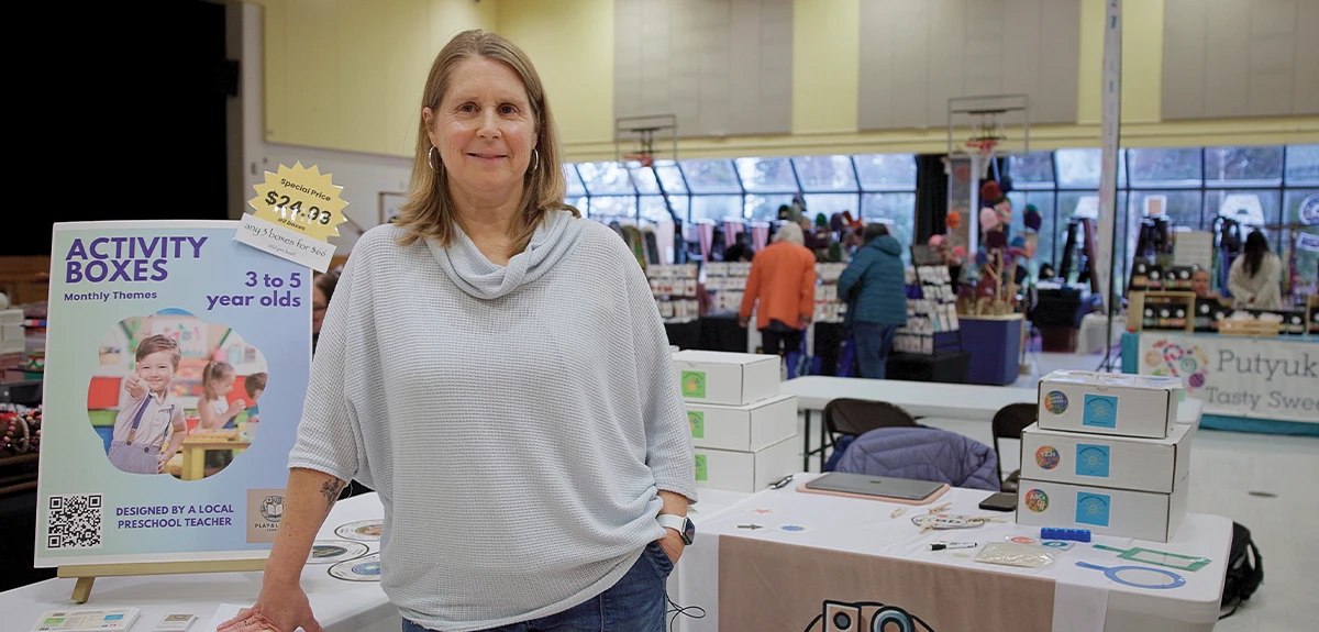 Kris Merizon, wearing a light gray sweater, stands smiling behind a vendor table at a market in a gymnasium. A sign for "Activity Boxes" and stacks of white subscription boxes are on the table in front of her.