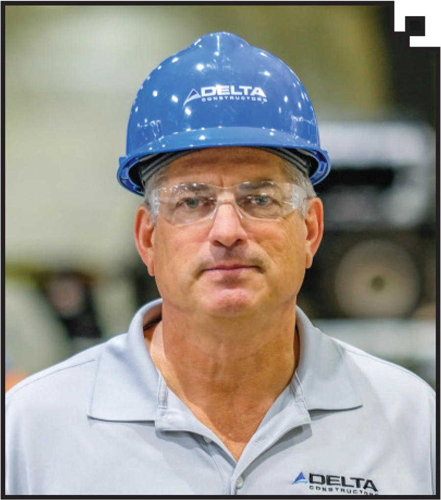 headshot of Ed Gohr in a factory wearing a blue hardhat labeled with the Delta Constructors logo