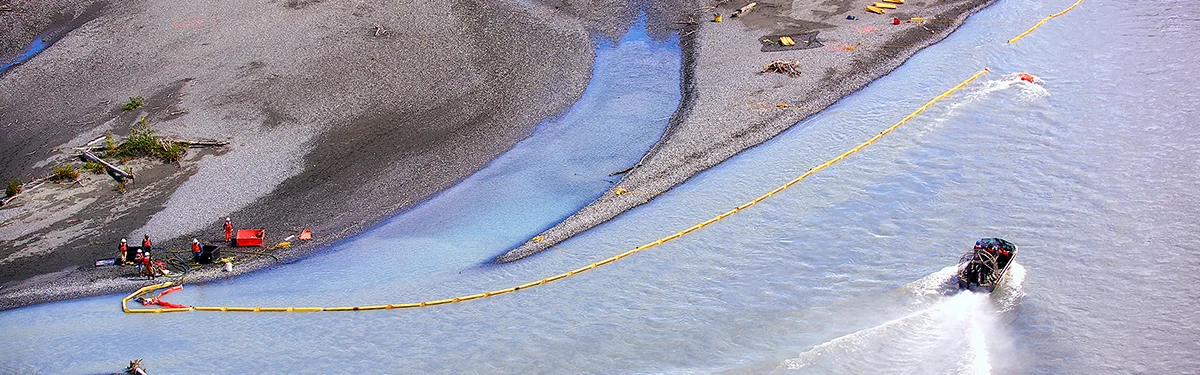 High-angle aerial view of a boat deploying a long yellow containment boom across silty water toward a gray shoreline. Crew members in orange safety gear stand near equipment on the gravel beach.