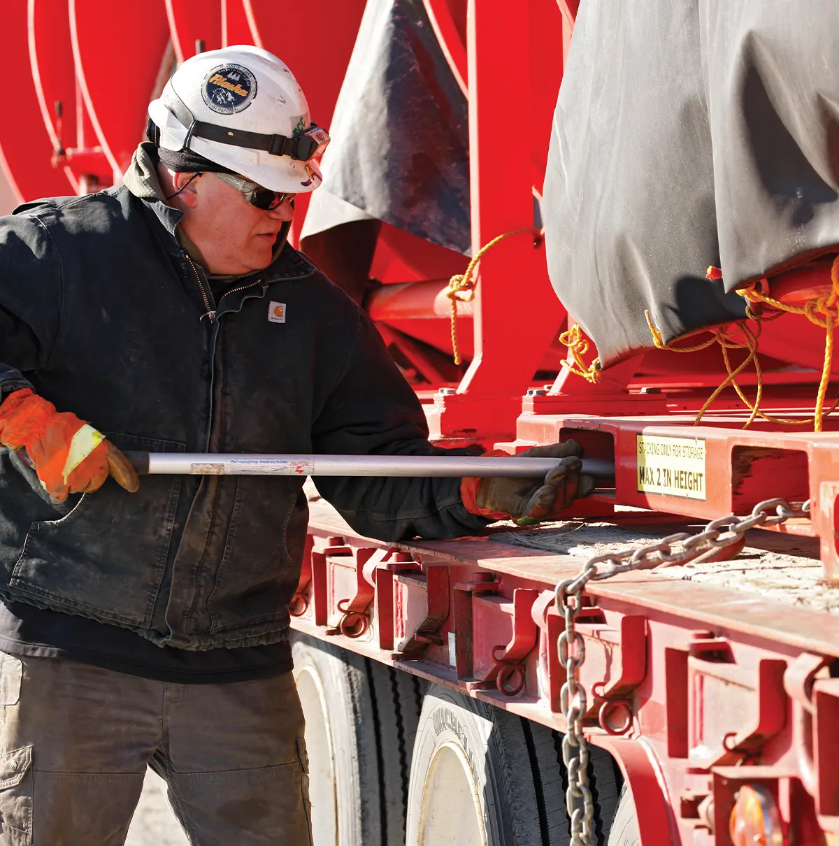 A worker in a black jacket, white hard hat, and orange gloves uses a metal lever to secure heavy equipment on a red flatbed trailer. Large red machinery is visible in the background.