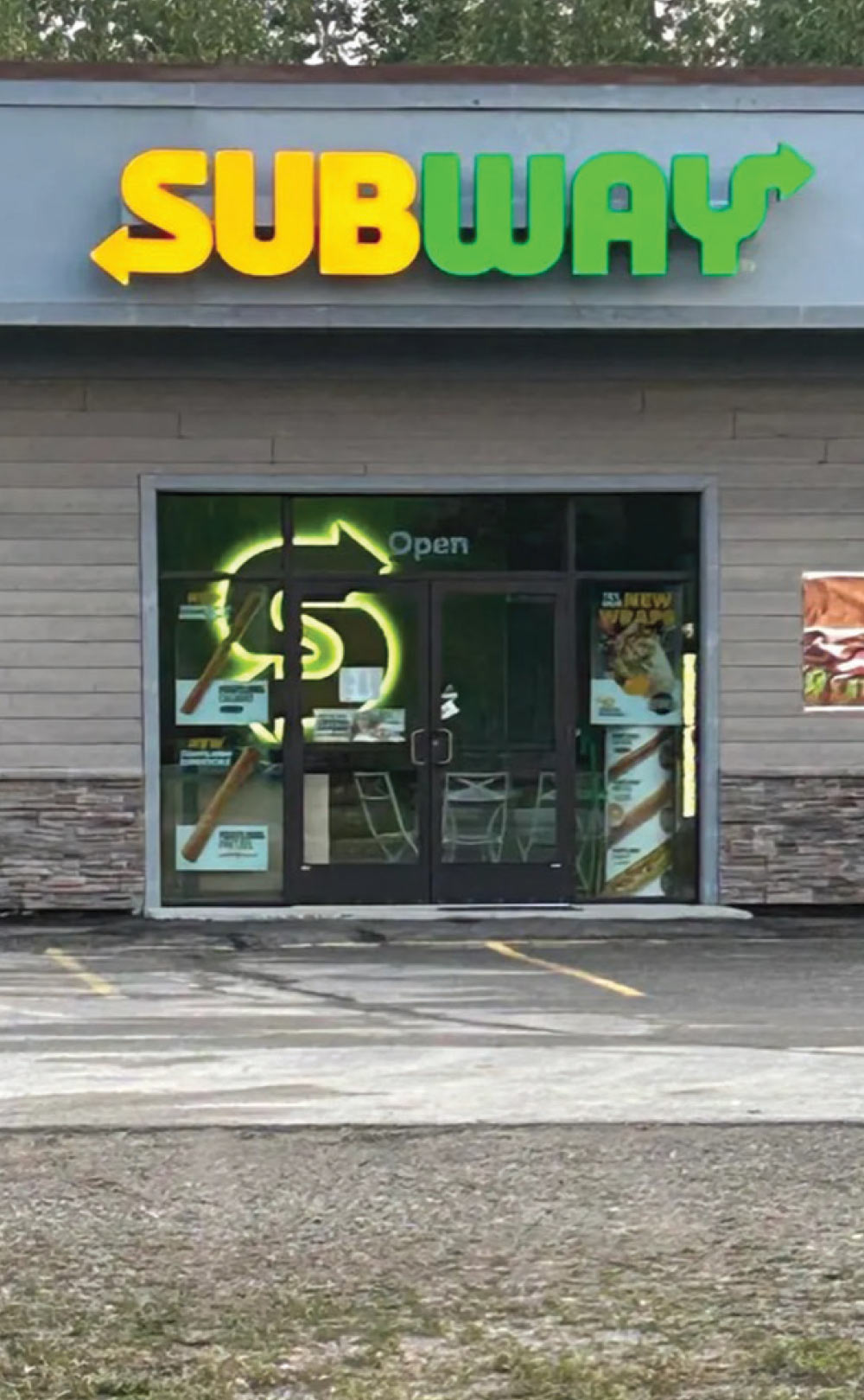 The exterior entrance of a Subway restaurant with a glowing neon "S" logo visible through the glass door.