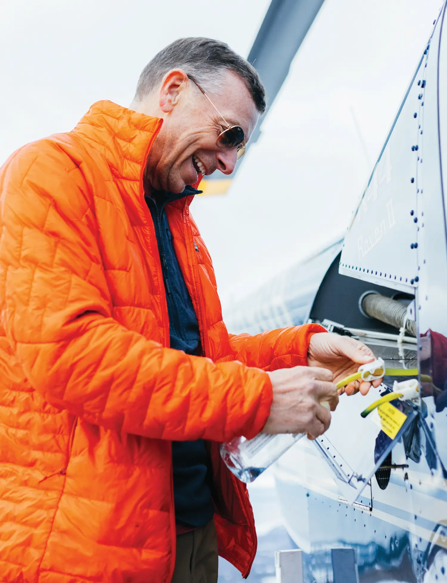 Sean McLaughlin, a smiling man wearing an orange Patagonia puffer jacket and sunglasses, is smiling while performing maintenance or an inspection on a blue helicopter; He appears to be handling a small component or sensor near an open access panel on the aircraft's fuselage