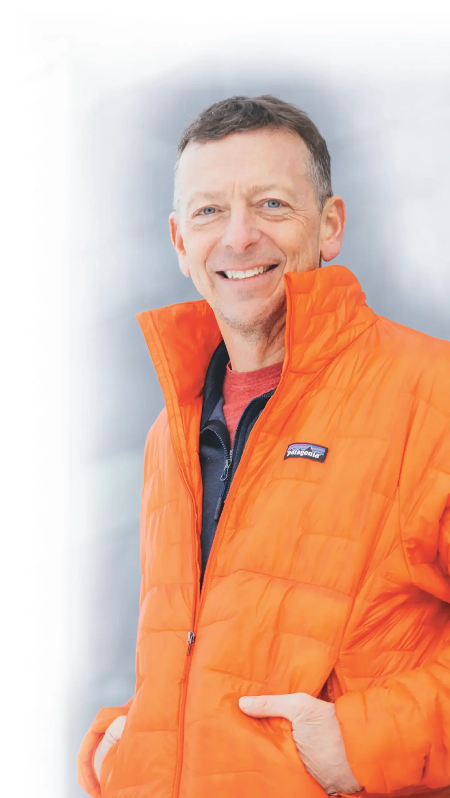 Sean McLaughlin, a smiling man wearing an orange Patagonia puffer jacket over a red shirt and a black zip-up, is seen standing with his hands inside the pockets of his puffer jacket