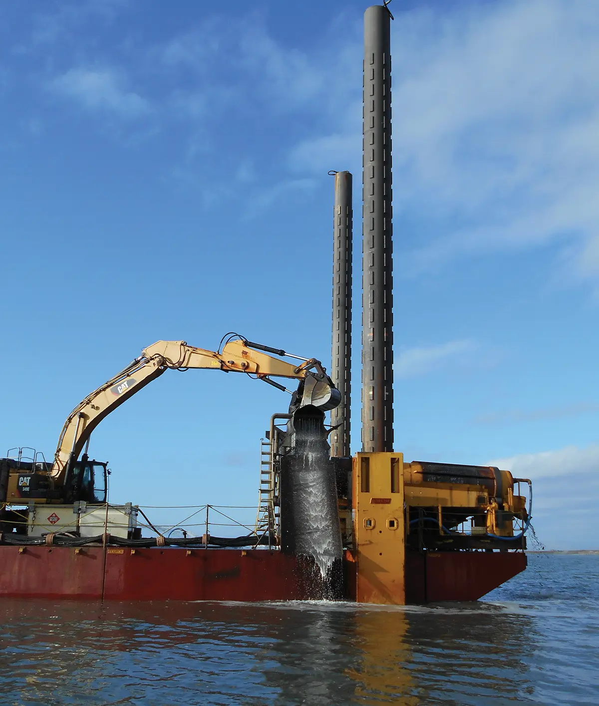 A yellow CAT excavator mounted on a red barge pouring sediment and water into a processing unit. Two tall, grey vertical spuds rise from the barge into a blue sky with light clouds. The vessel sits on open water.