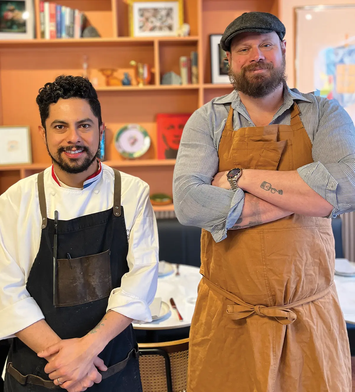 Two men stand in a restaurant. The man on the left wears a white chef's coat and black apron. The man on the right wears a flat cap and tan apron with arms crossed. Behind them are orange shelves with various books and decor.