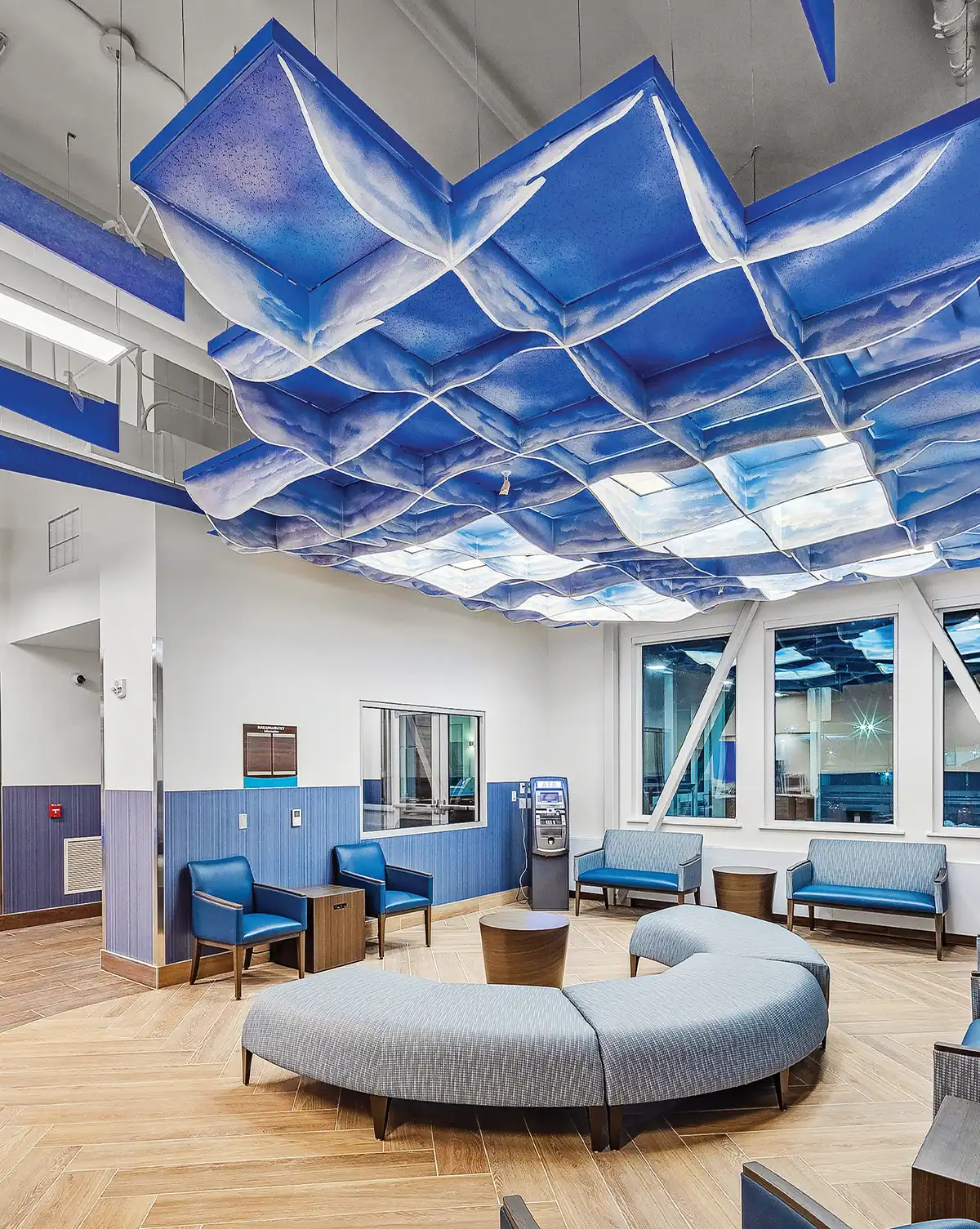 A modern lobby featuring blue seating and a curved ottoman on herringbone floors. A blue and white wavy grid installation hangs from the ceiling. The room includes blue wainscoted walls, large windows, and an automated kiosk.