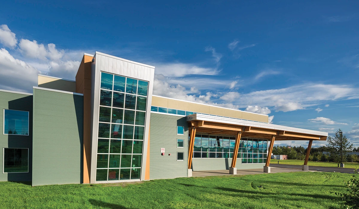Modern building with green corrugated siding, tan accents, and a large angled glass grid window. A wooden-beamed canopy extends over the walkway. Set on a green lawn under a bright blue sky with wispy clouds.