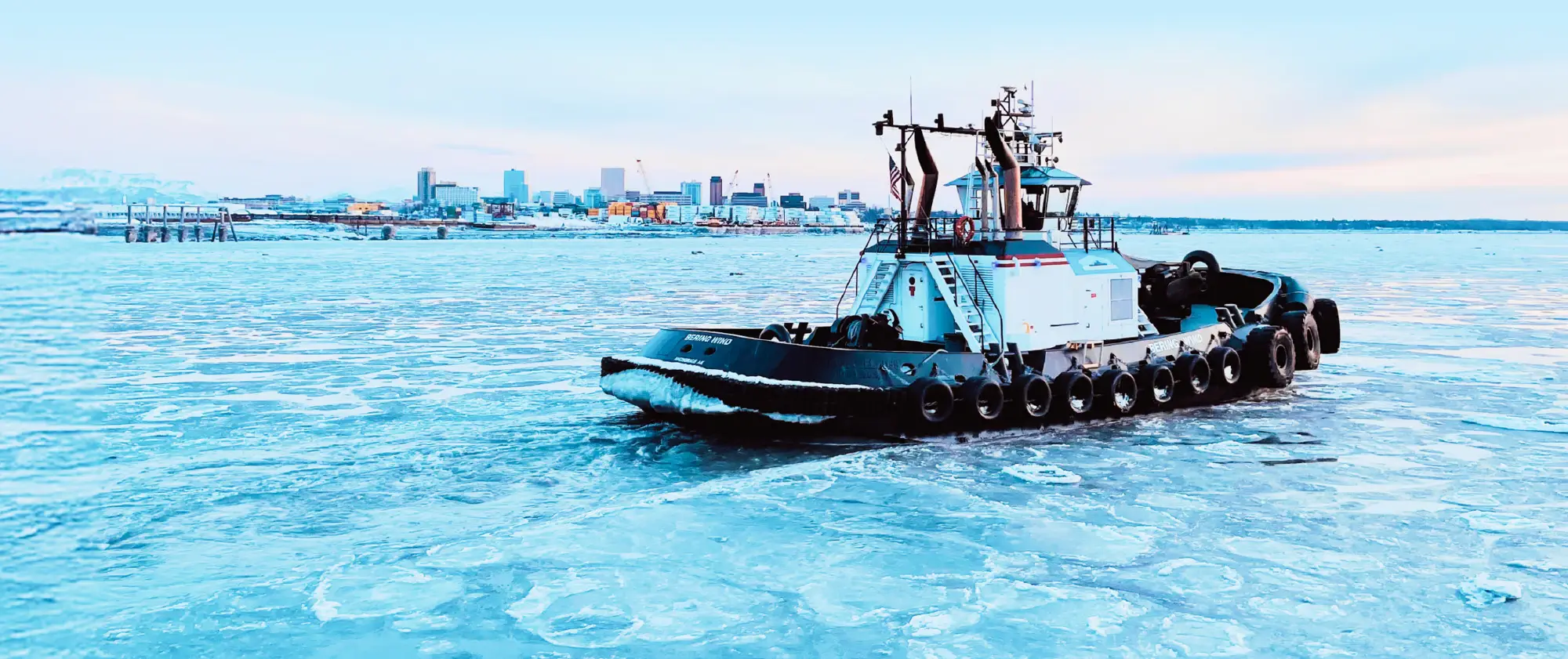 Industrial tugboat equipped with heavy-duty tires for fendering, pushing through frozen waters during a cold winter day in Alaska.