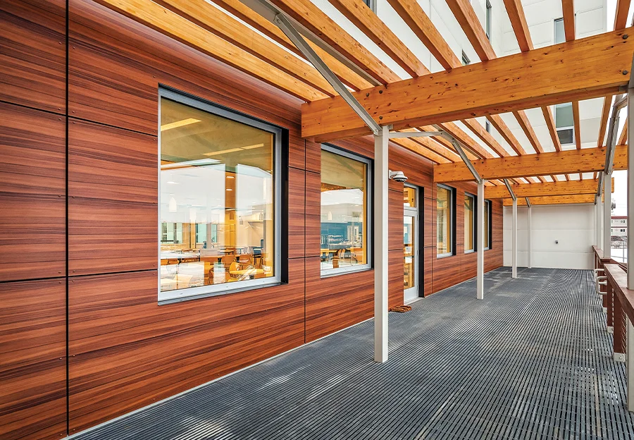Close-up of the Qavartarvik Customer Lodging exterior featuring rich wood-look wall panels, a heavy timber pergola structure, and industrial metal grate flooring under a covered walkway
