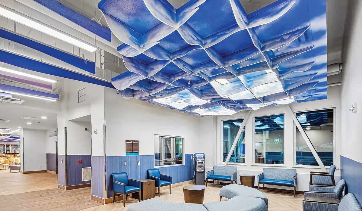 Interior of a modern hospital waiting room featuring a dramatic blue cloud-patterned sculptural ceiling installation, blue upholstered seating, and large windows with integrated structural bracing