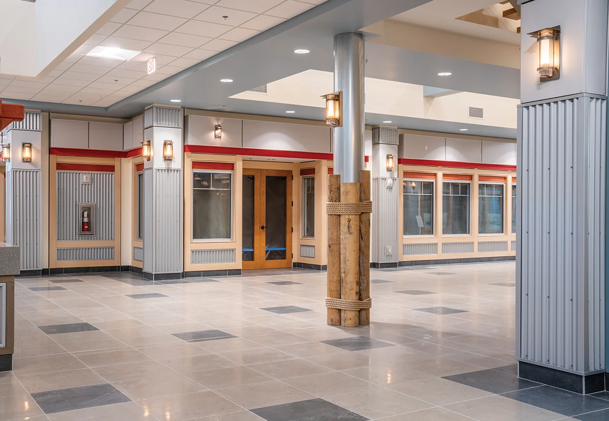 Interior hallway of a modern commercial facility featuring polished gray tile flooring, corrugated metal wall accents, and a unique structural pillar wrapped in decorative wood logs and rope
