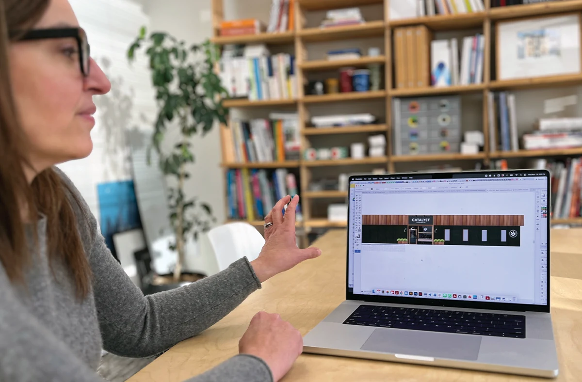 Close-up of a professional at a wooden desk using design software to visualize a retail exterior, with a large plant and bookshelf in the background.