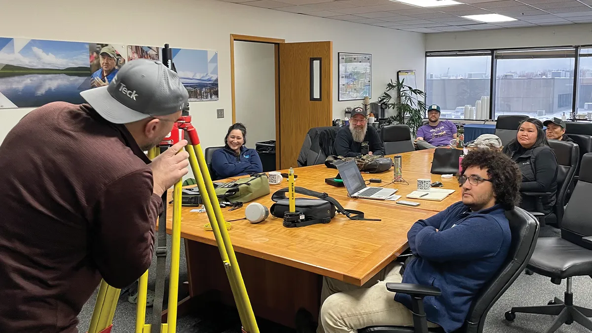 Interior view of an Alaska Business office meeting where a field technician demonstrates surveying tools to a group of seated colleagues
