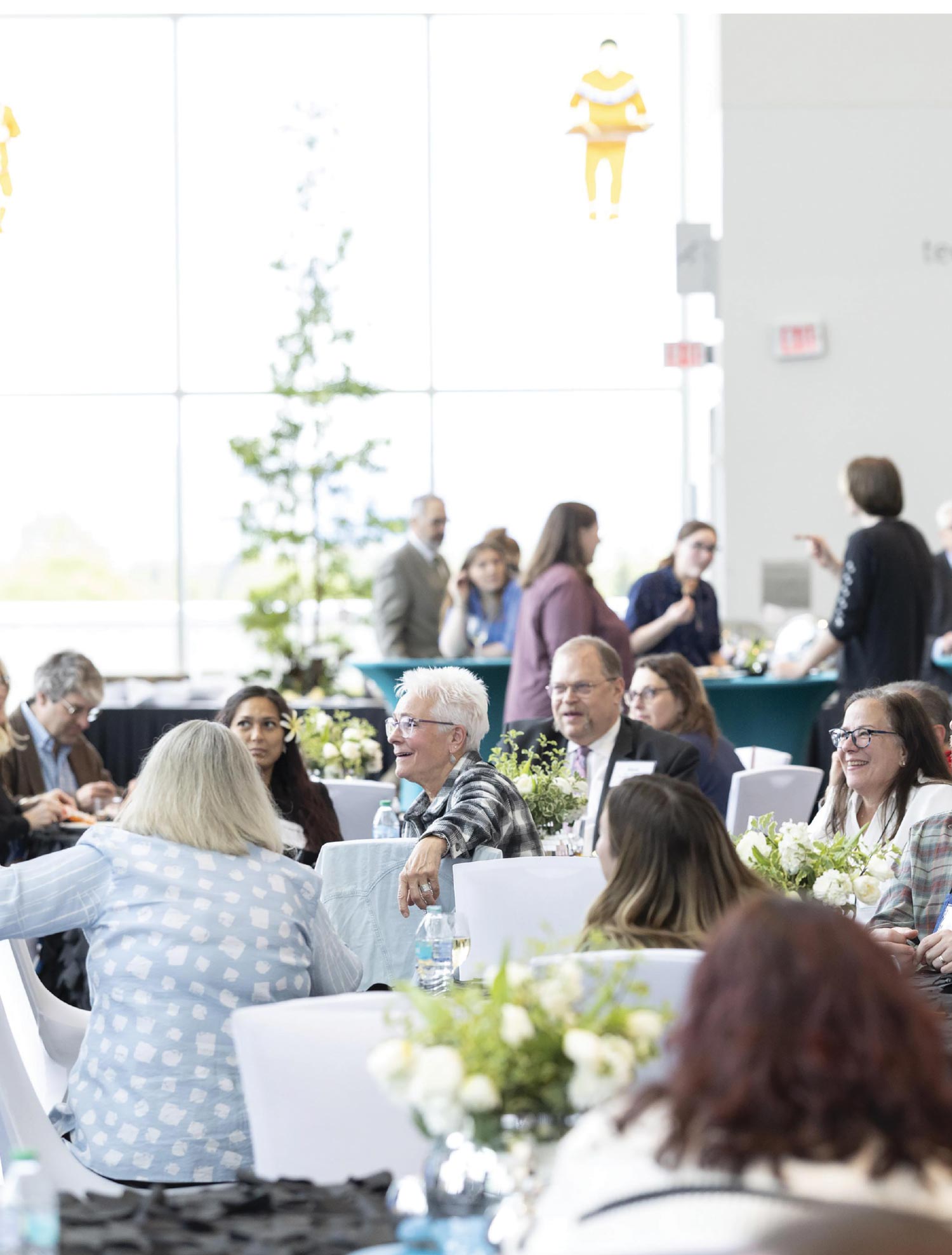 People gathered around round tables in a bright, high-ceilinged event space with a tall vertical plant arrangement.