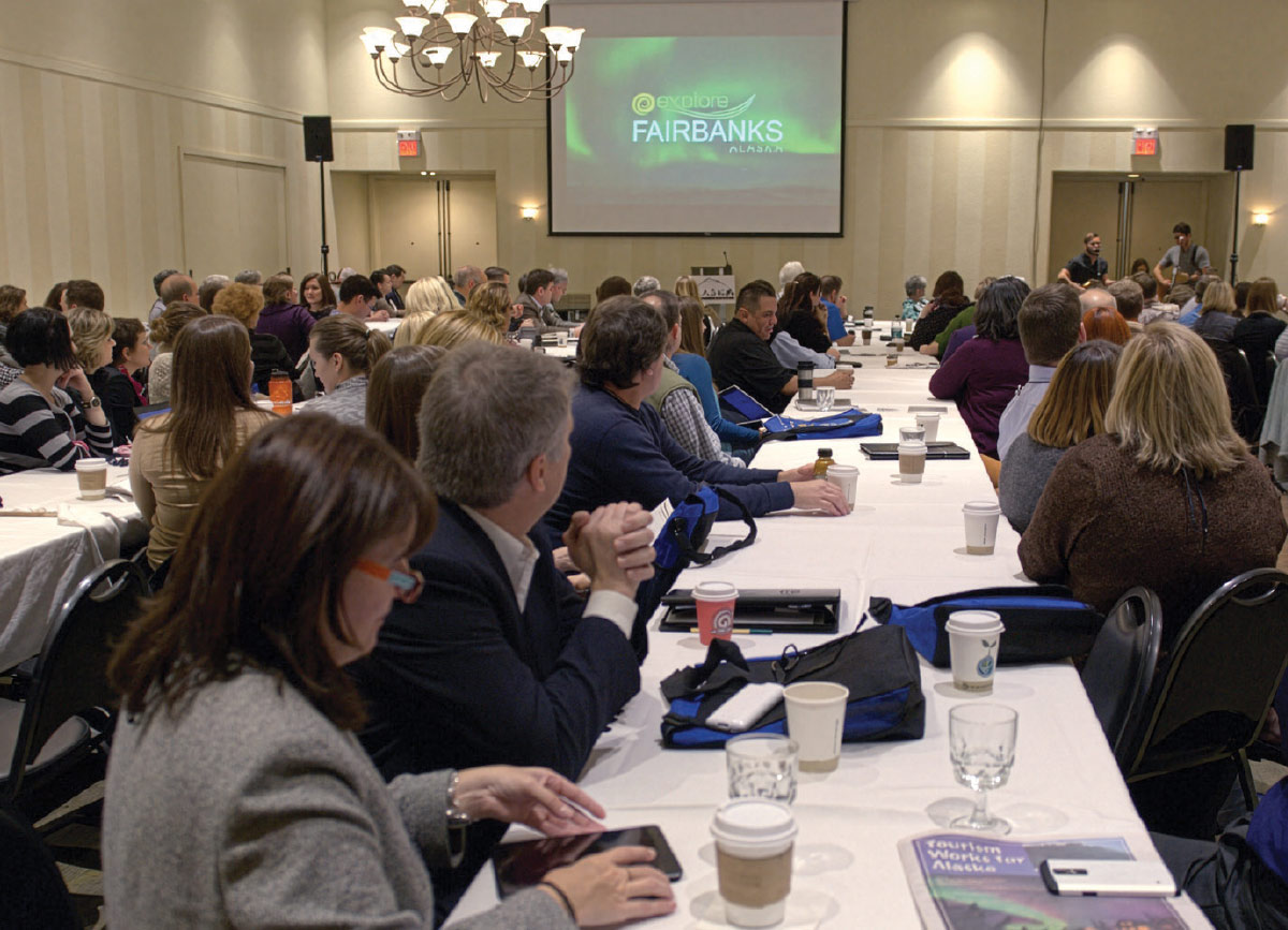 Professionals sit at long tables in a conference room, facing a screen displaying "explore FAIRBANKS ALASKA."
