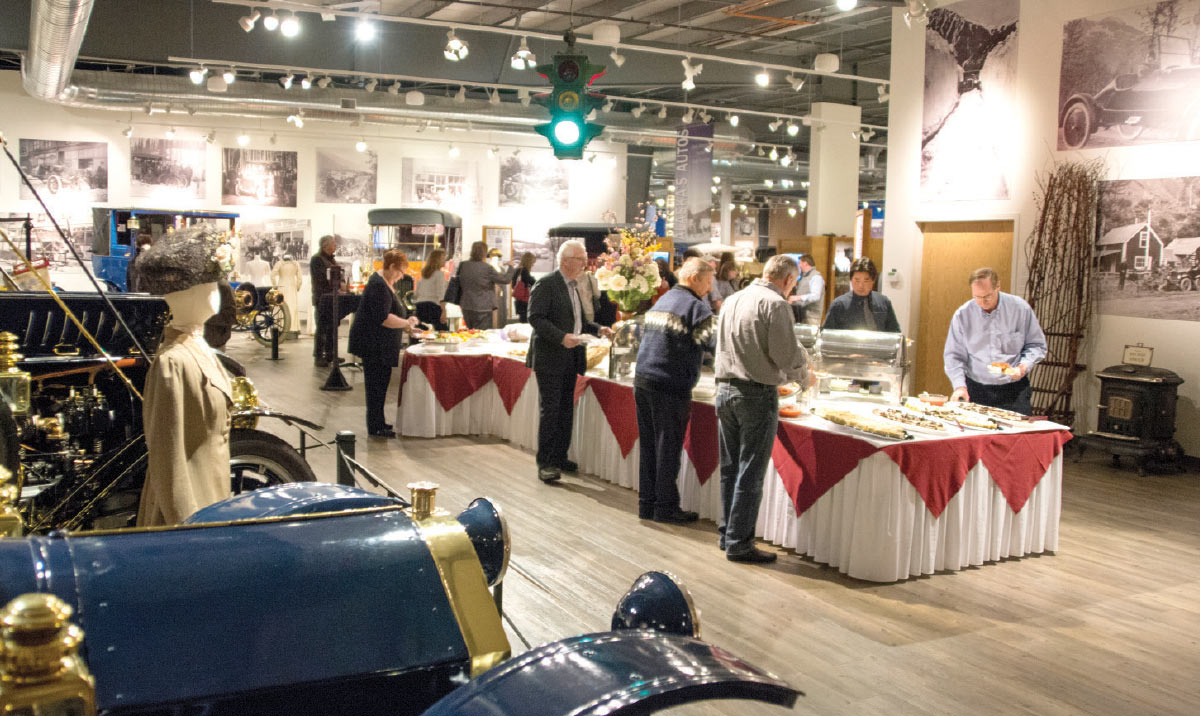 People serving themselves from a buffet line set up inside an auto museum featuring vintage blue cars.