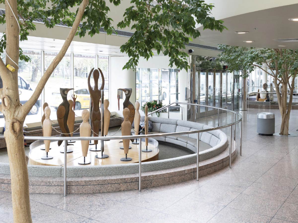 Indoor garden area with wooden bird and human-shaped sculptures surrounded by a circular bench and indoor trees.