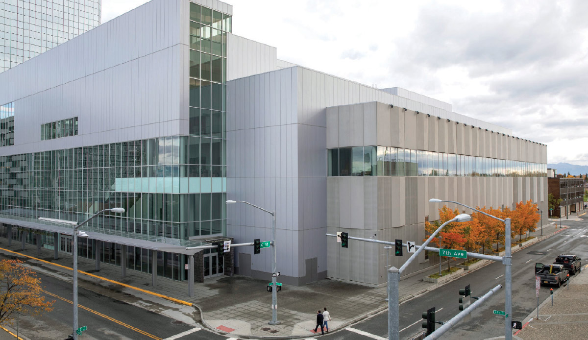 High-angle exterior view of a modern multi-story glass and grey building at a city street corner with autumn trees.