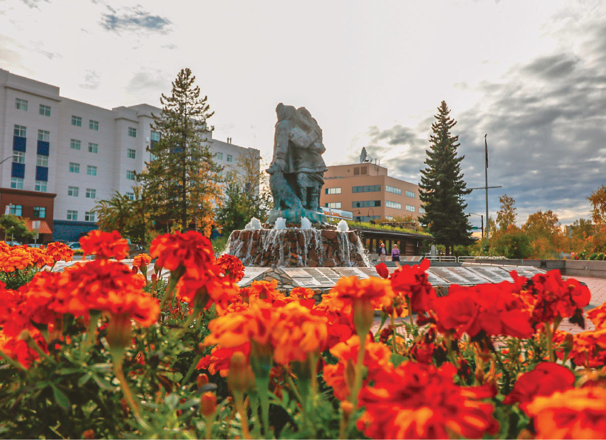 A stone fountain with a sculpture of two figures stands behind a flowerbed of bright orange marigolds.