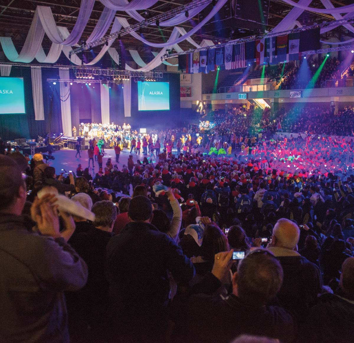 Wide shot of a massive crowd in an arena watching a performance on a stage with large "ALASKA" video screens.