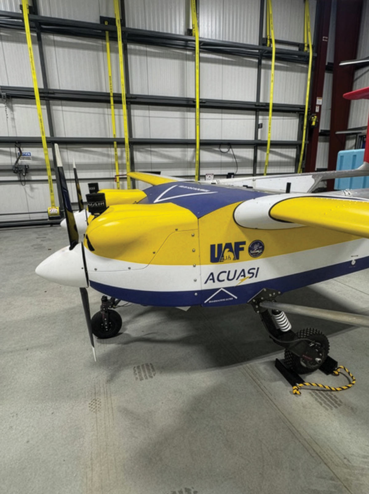 Close-up of a yellow, white, and blue fixed-wing drone with "UAF ACUASI" branding and twin propellers in a hangar.