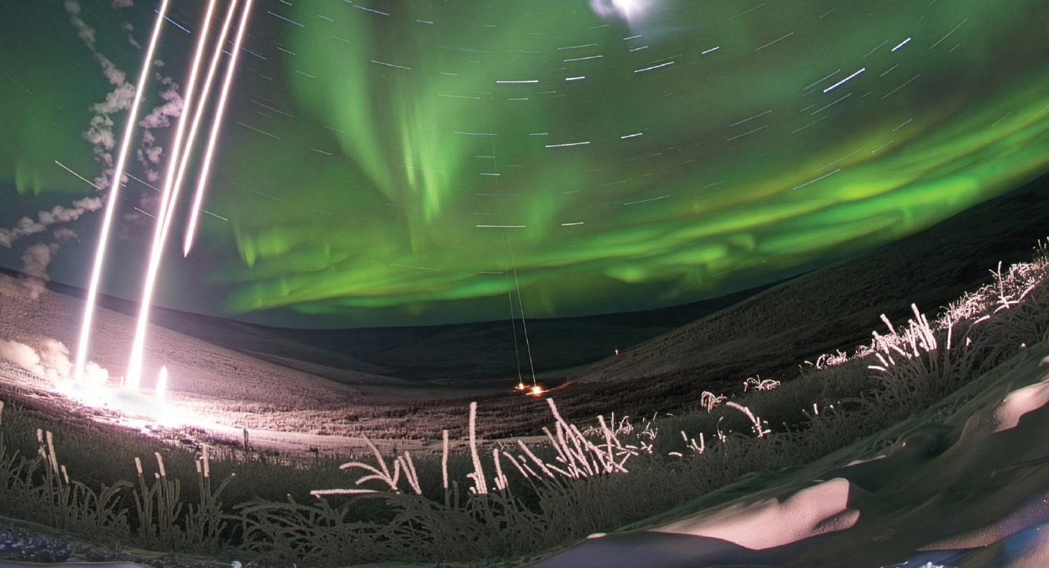 Long-exposure night shot of four rockets launching into a bright green aurora borealis over a snowy Alaskan landscape.