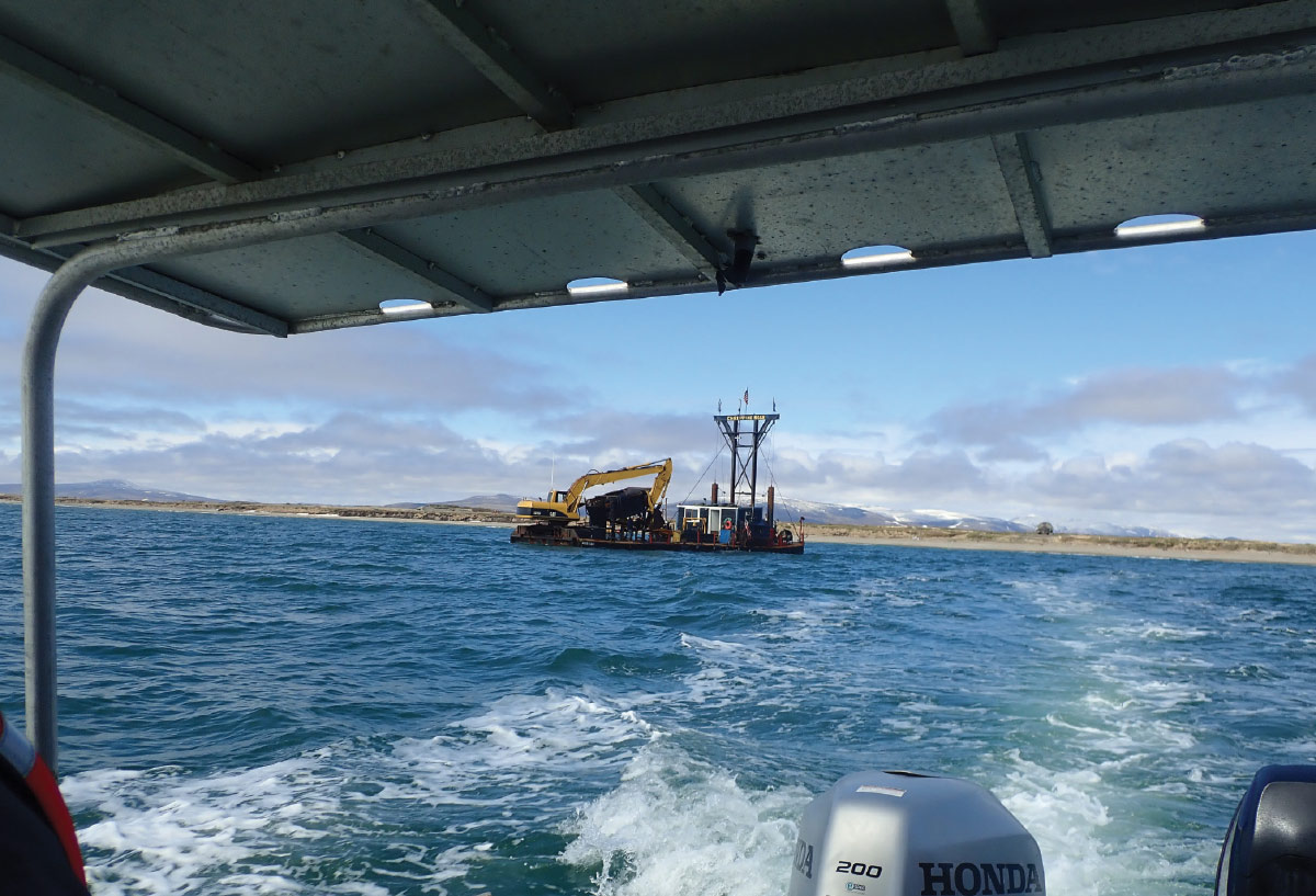 View from a moving boat showing a gold dredge with a yellow excavator operating in open water near a coastline.