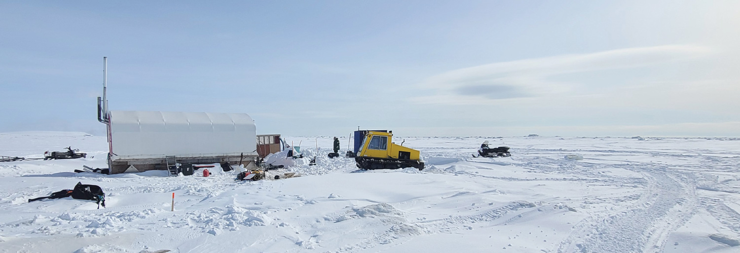 A wide, snowy landscape featuring a yellow tracked vehicle, a snowmobile, and a white temporary shelter on the ice.