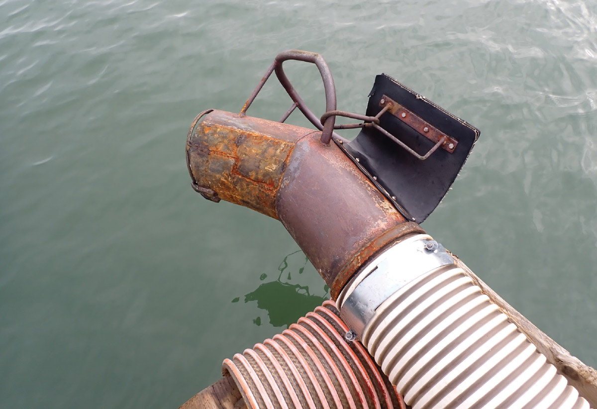 Close-up of a rusted metal discharge pipe and flexible industrial hoses extending over open water.