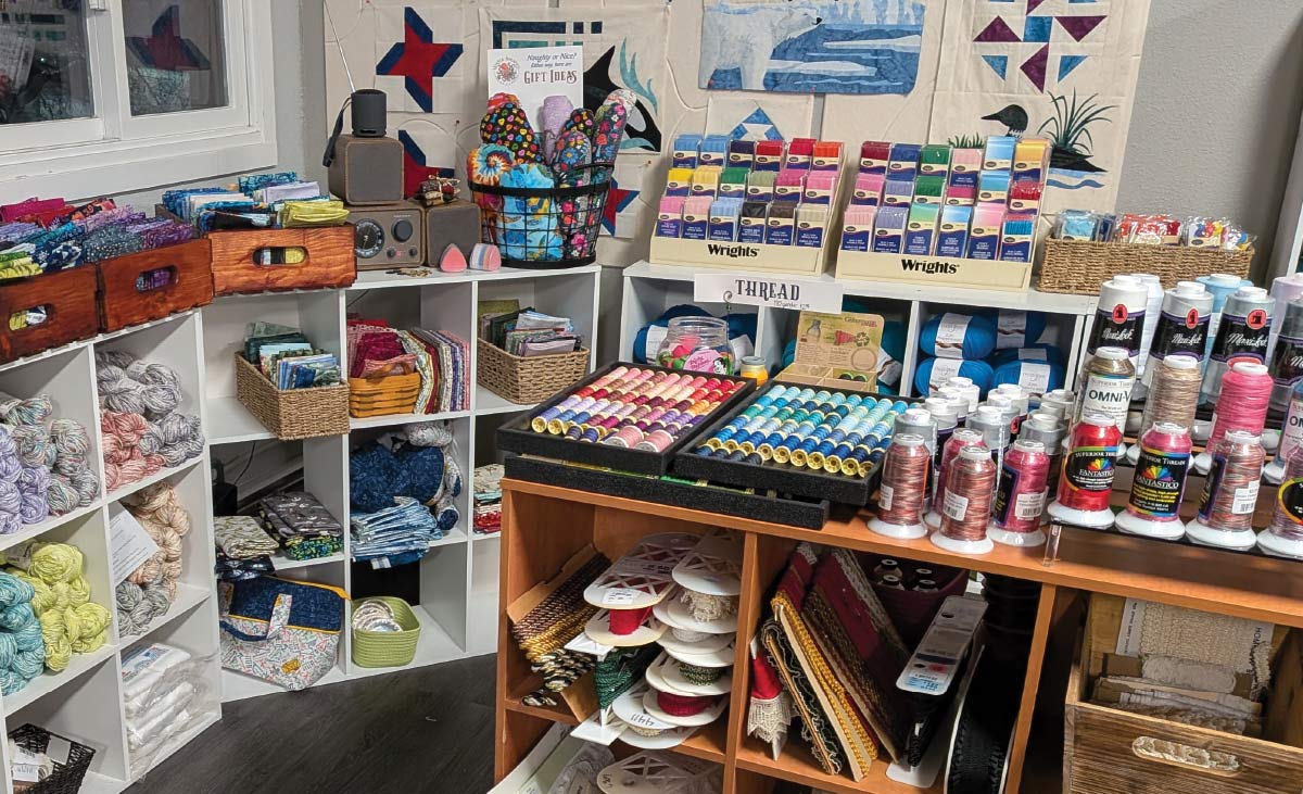 Craft store display featuring shelves of yarn, baskets of colorful thread spools, and various sewing notions on wooden tables.