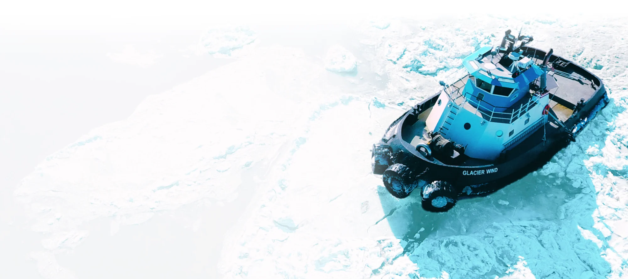 Aerial view of the "Glacier Wind" tugboat navigating through thick, broken ice floes in a frozen Alaskan harbor.