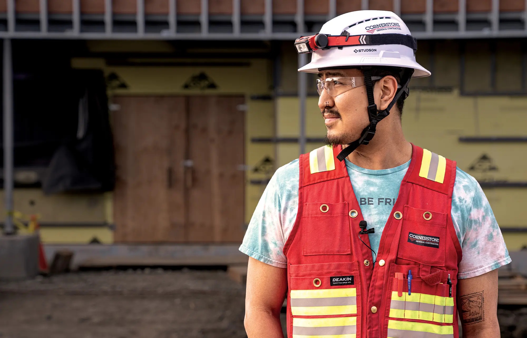 A construction site manager wearing a white Cornerstone General Contractors hard hat, safety glasses, and a red high-visibility reflective vest.