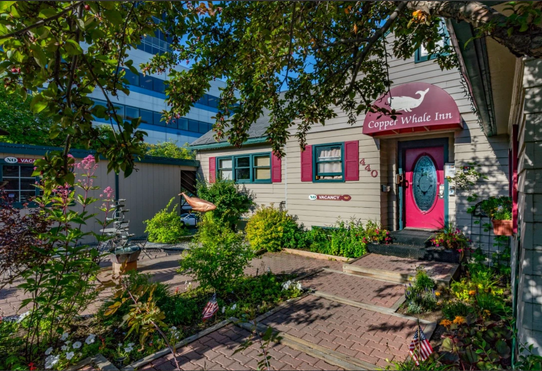 Exterior view of the Copper Whale Inn showing the front entrance, garden landscaping, and red door beneath the inn’s whale logo sign.