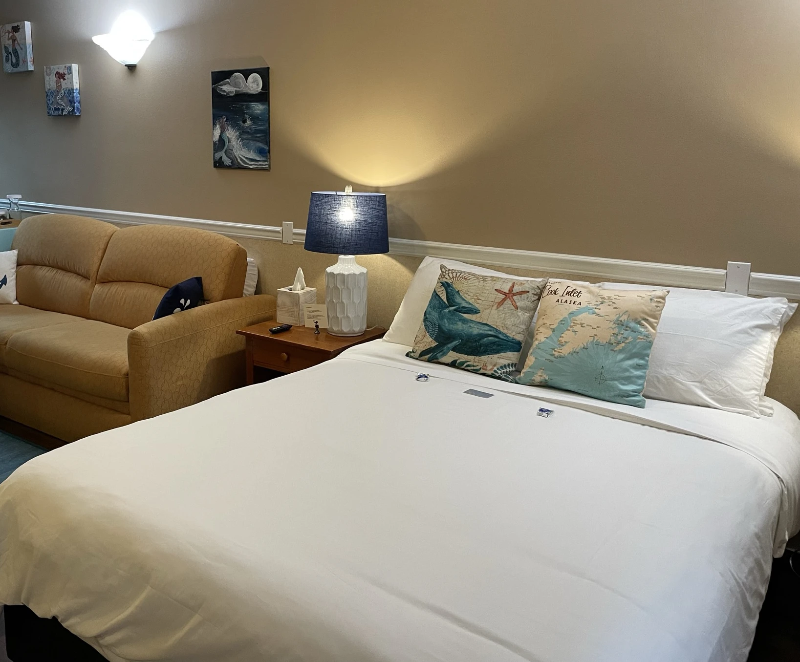 Guest room interior featuring a neatly made bed with whale-themed pillows, bedside table with lamp, and a sofa against a warm beige wall.