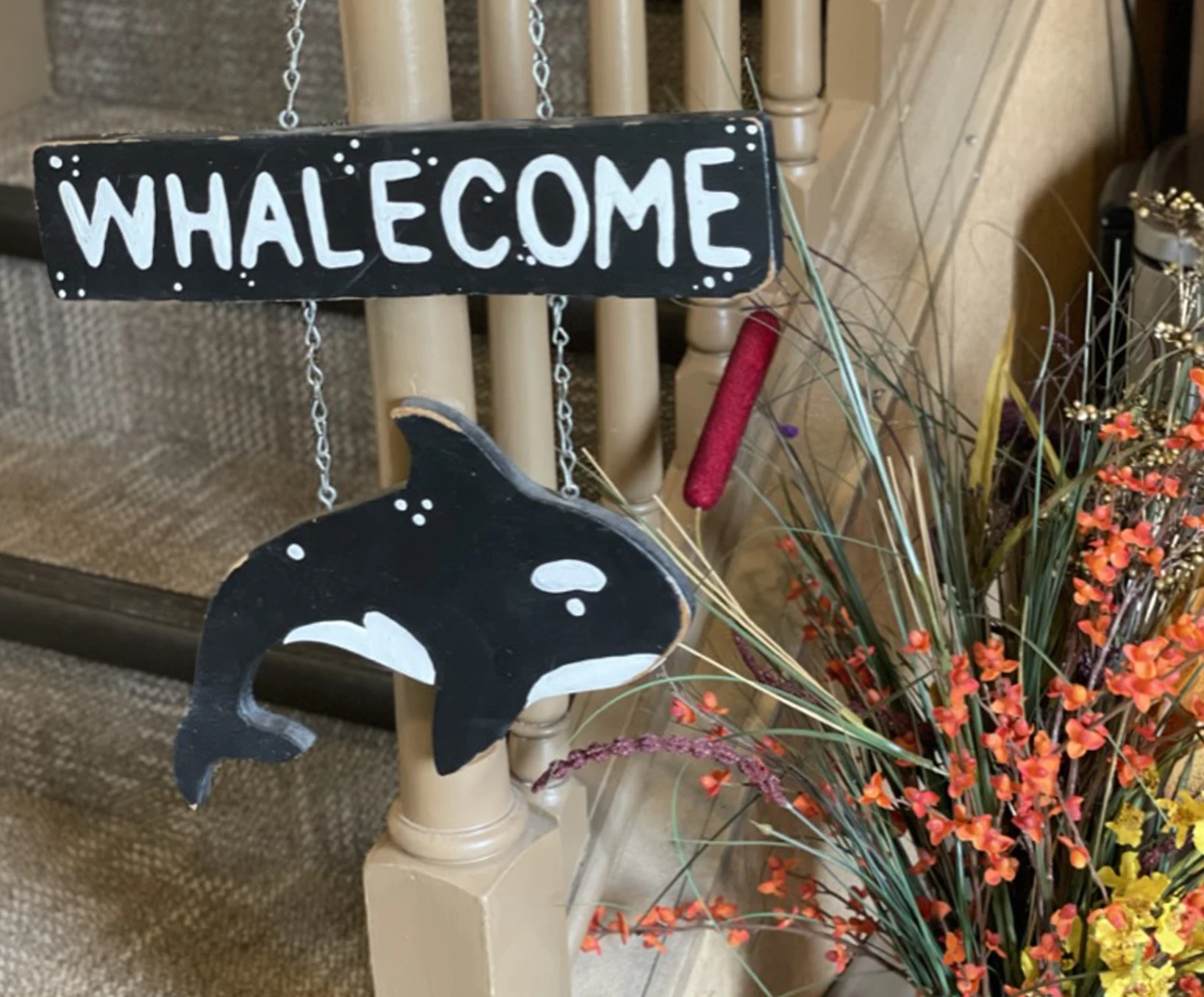 Decorative wooden sign reading “Whalecome” with a painted orca hanging below it, displayed near a staircase with floral decor.