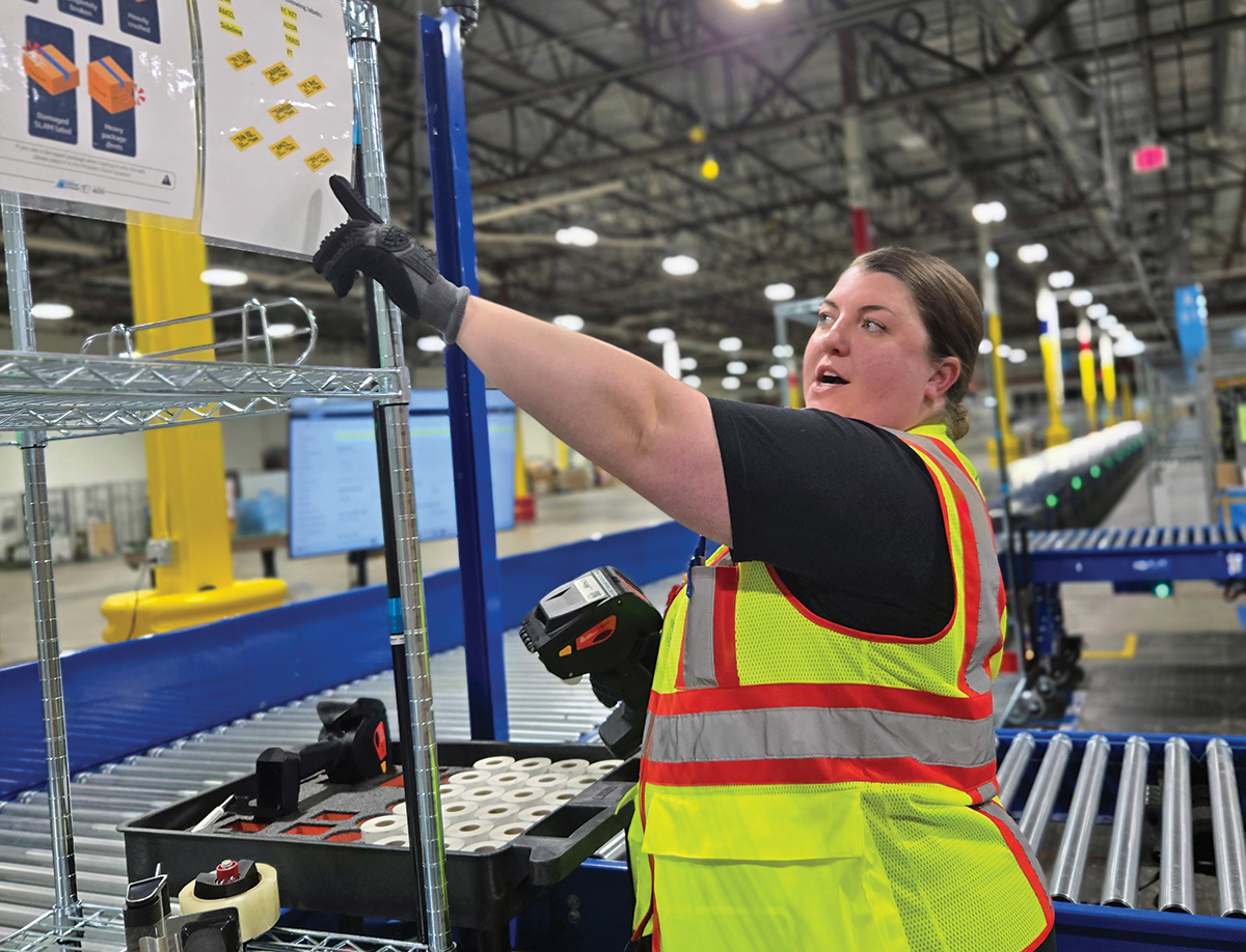 WFB1 Site Leader Cydney Edge, a woman in a yellow safety vest, points toward an instructional sign mounted on a silver wire shelving unit in a warehouse; A blue conveyor belt and several rollers are visible in the background