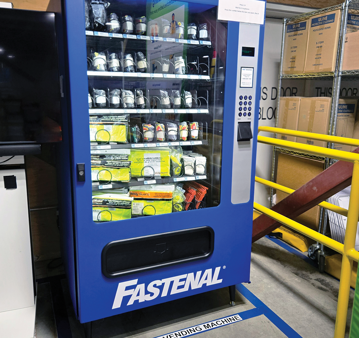 A large, bright-blue FASTENAL industrial vending machine sits on a warehouse floor; The machine's glass front reveals rows of organized safety equipment, including gloves, high-visibility vests, and other protective gear