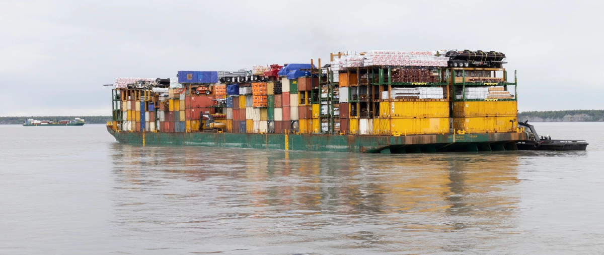 A wide shot of a large, green-hulled transport barge heavily loaded with a diverse array of multi-colored shipping containers and lumber, moving across a calm body of grey water