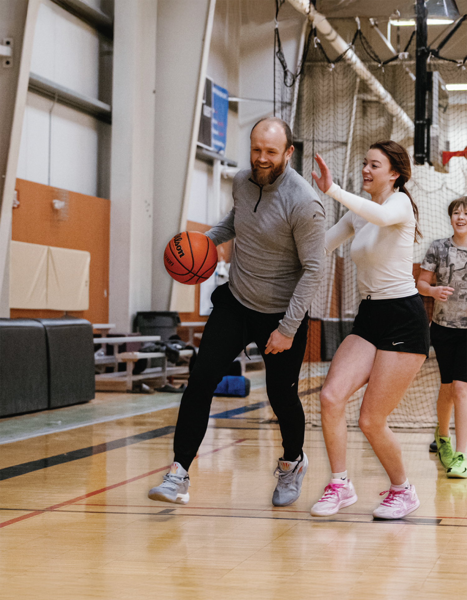 Buddy Bailey, a bald man with a dark brown beard facial hair smiles in a grey long-sleeve quarter-zip athletic shirt, black jogger pants, and grey fitness shoes as he is dribbling a basketball past a young female defender on a hardwood court; A younger boy watches from the background behind them