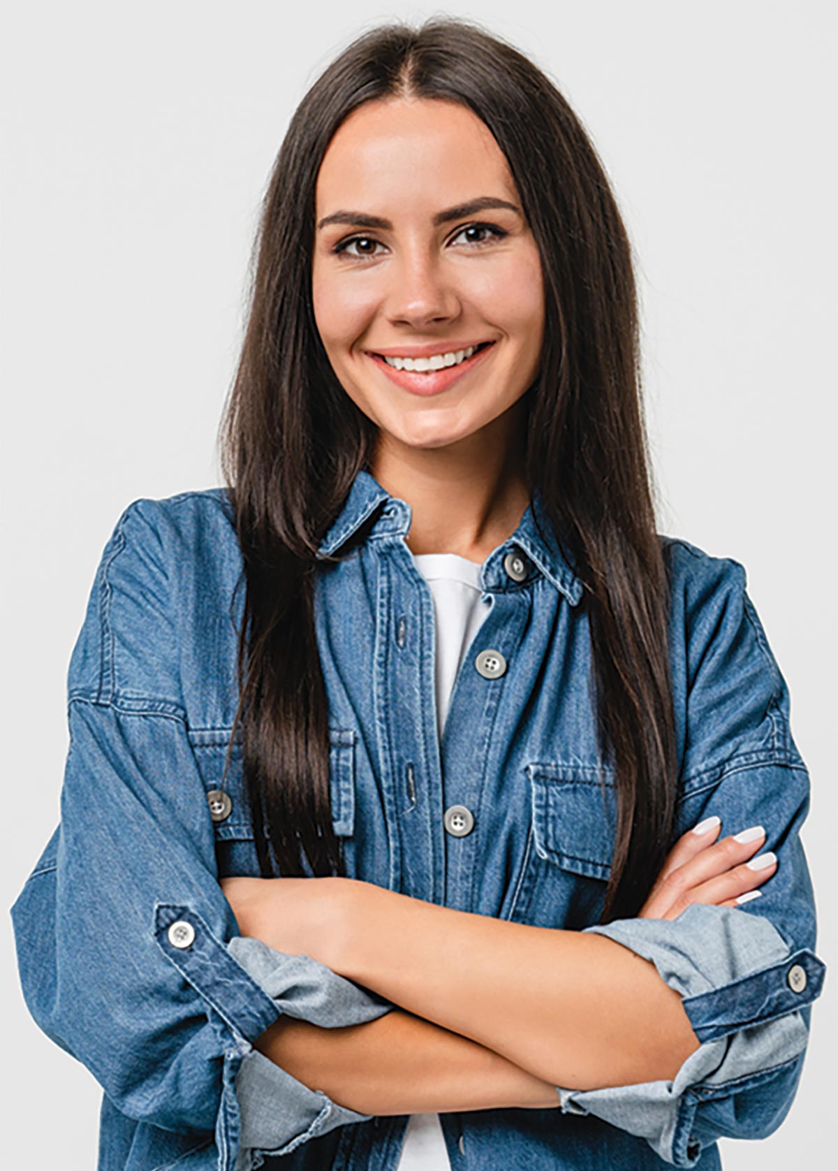A smiling woman with long dark hair wearing a denim shirt over a white t-shirt, standing with her arms crossed.
